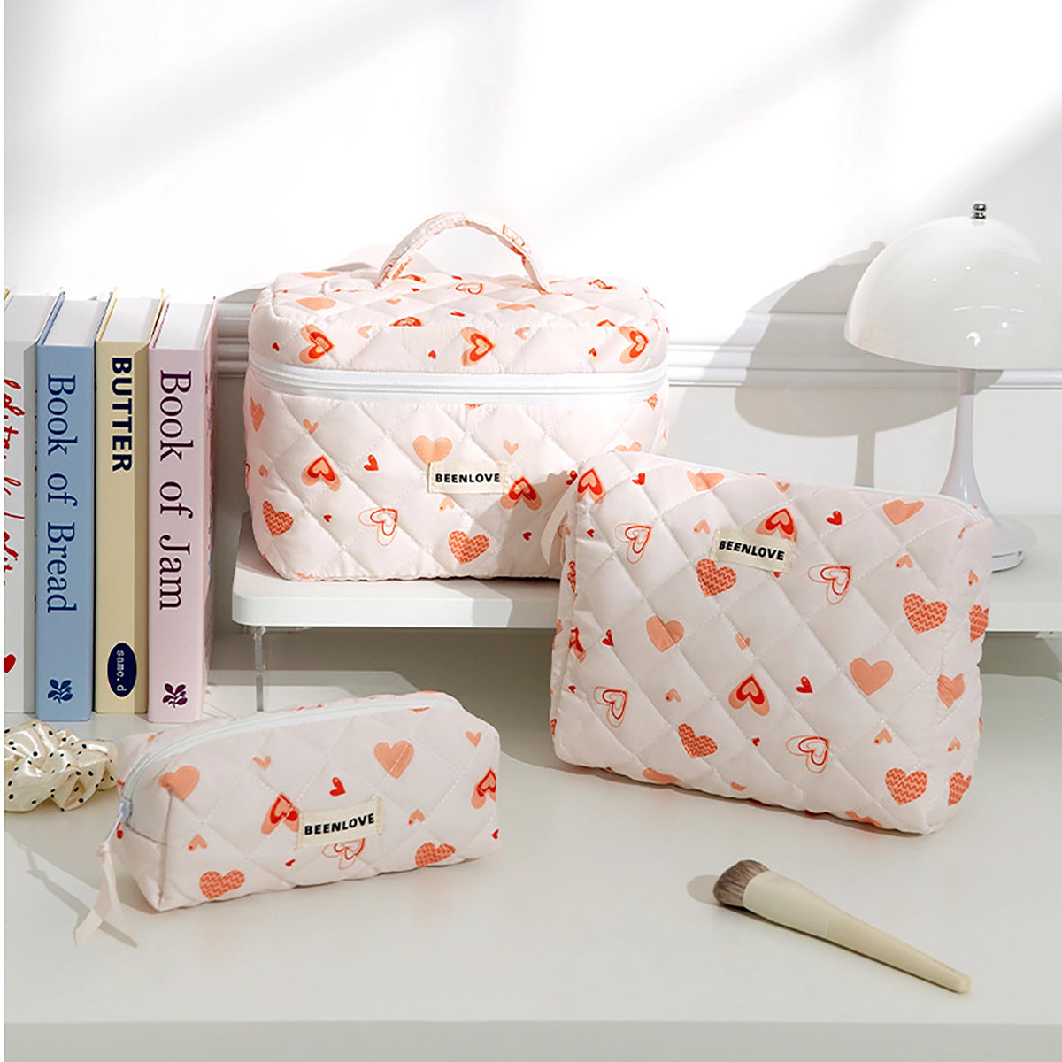 Three quilted cosmetic pouches in a soft blush pink fabric with an allover red and orange heart pattern, displayed on a white shelf and tabletop next to decorative books and a white lamp.
