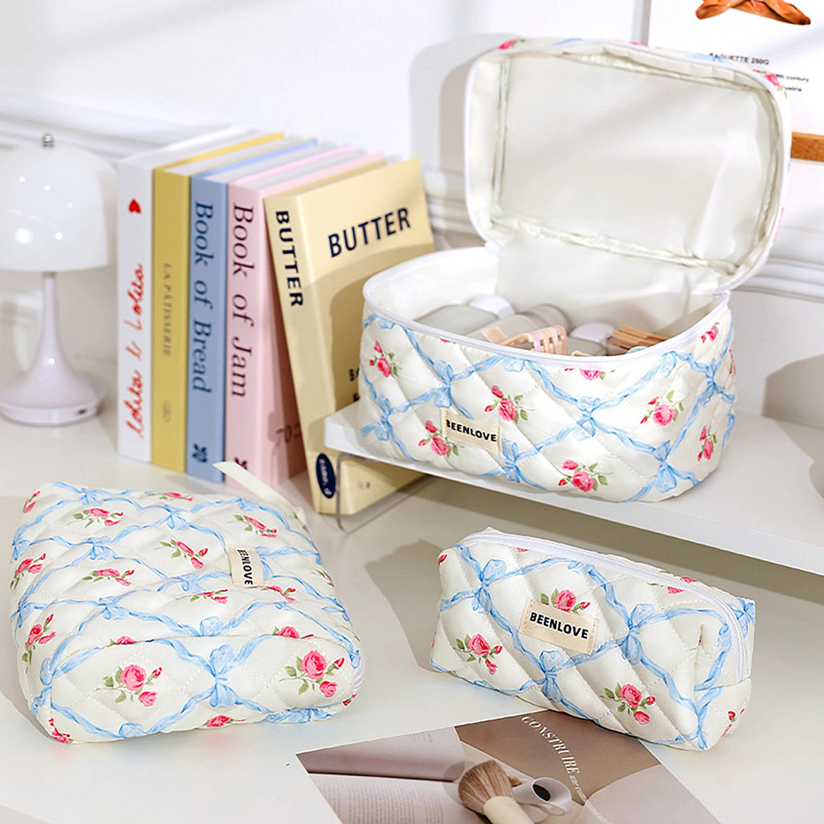 Three quilted cosmetic pouches with a cream floral print and blue lattice pattern are arranged on a white tabletop. One pouch is open, showing storage space inside, while the other two are closed.