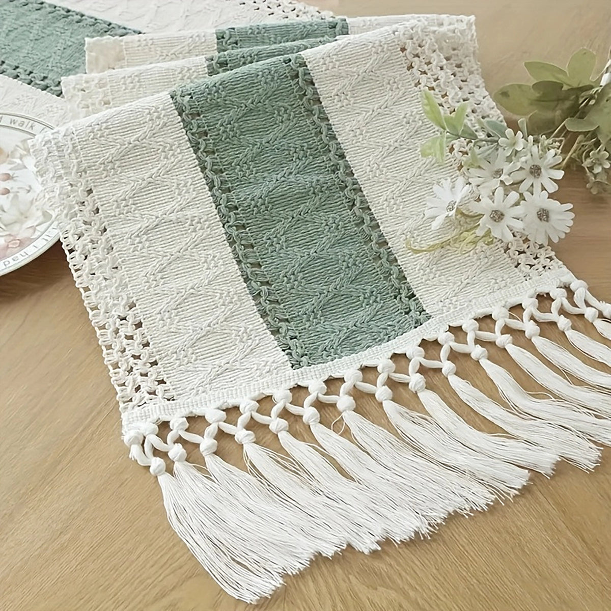 A rectangular lace table runner with fringe tassels, featuring a central sage green stripe flanked by white lace panels, displayed on a light wood tabletop with small white flowers.