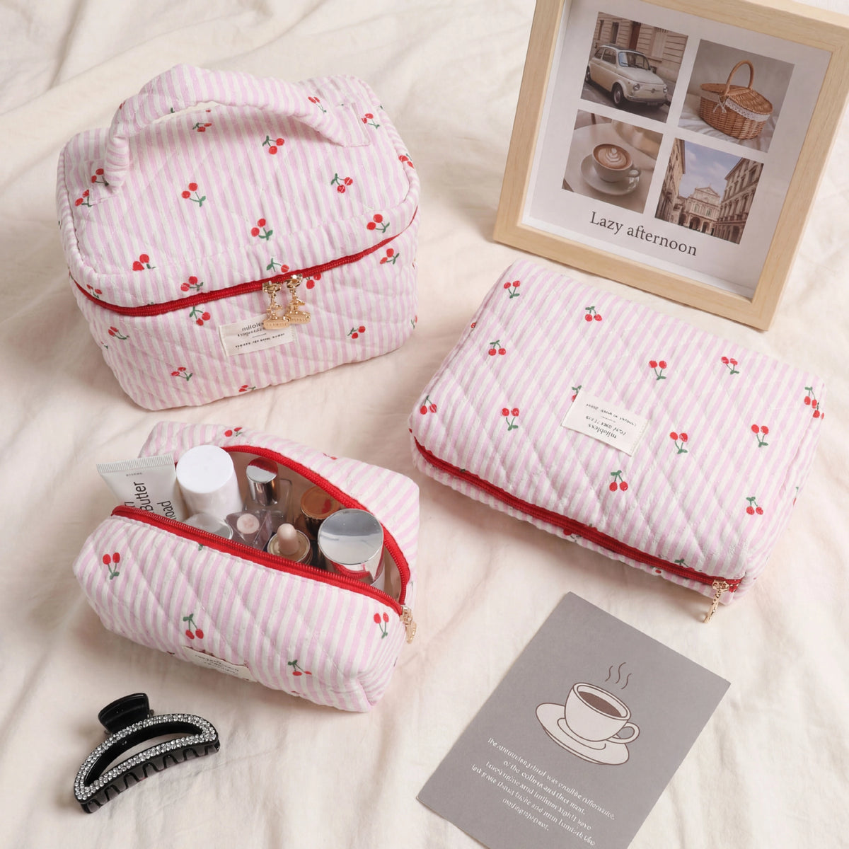 Three quilted pink cosmetic bags with a cherry print and red zipper trim are arranged on a cream fabric surface. One bag is open, showing makeup items inside, and a small black hair claw clip and a gray card are placed nearby.