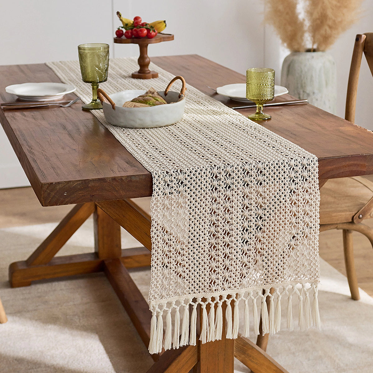 Wooden dining table with a beige crocheted table runner, green glasses, and fruit on a white wall background.