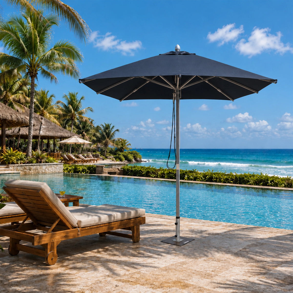 A large black patio umbrella stands beside a resort-style swimming pool on a stone deck, with a wooden lounge chair in the foreground and palm trees, beach, and ocean in the background.