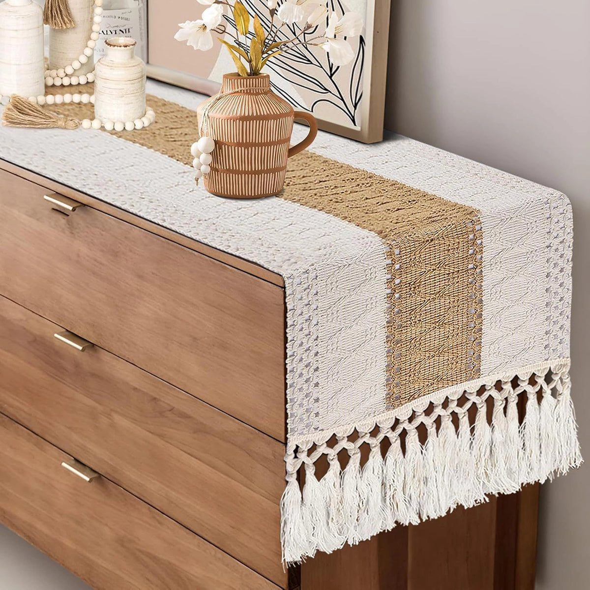 A woven table runner with a cream and natural jute stripe design and white fringe tassels, styled on top of a wooden dresser in a home decor setting.