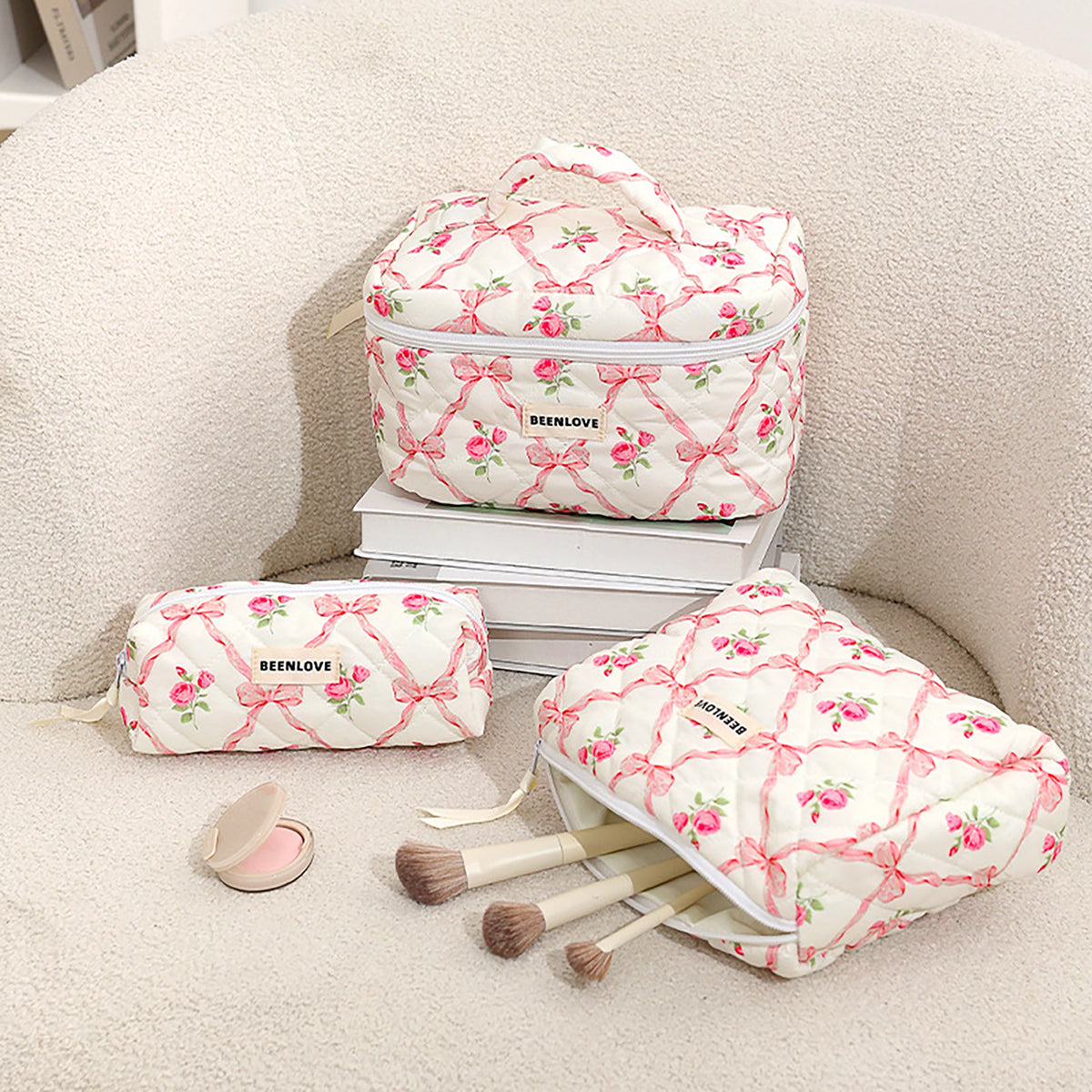 Three quilted makeup bags with a pink bow and floral print are arranged on a cream-colored textured chair. One large bag sits on stacked books, a smaller matching bag rests beside it, and an open bag lies on its side with makeup brushes and a compact powder inside.